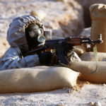 A Syrian soldier aims an AK-47 assault rifle from his position in a foxhole during a firepower demonstration, part of Operation Desert Shield.  The soldier is wearing a Soviet-made Model ShMS nuclear-biological-chemical warfare masks.